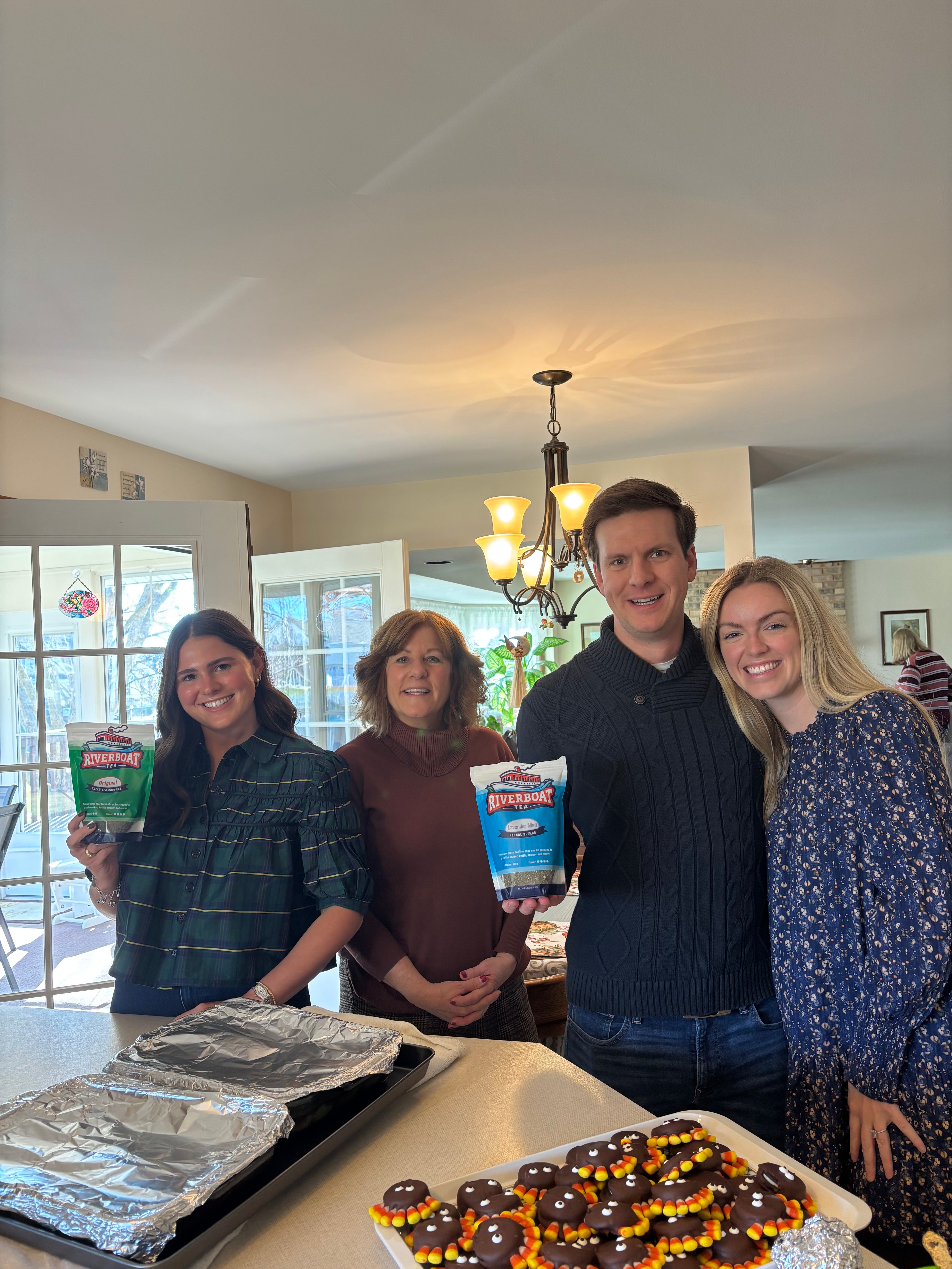 Group of people standing together in a room with a table full of snacks while holding pouches of Riverboat Tea